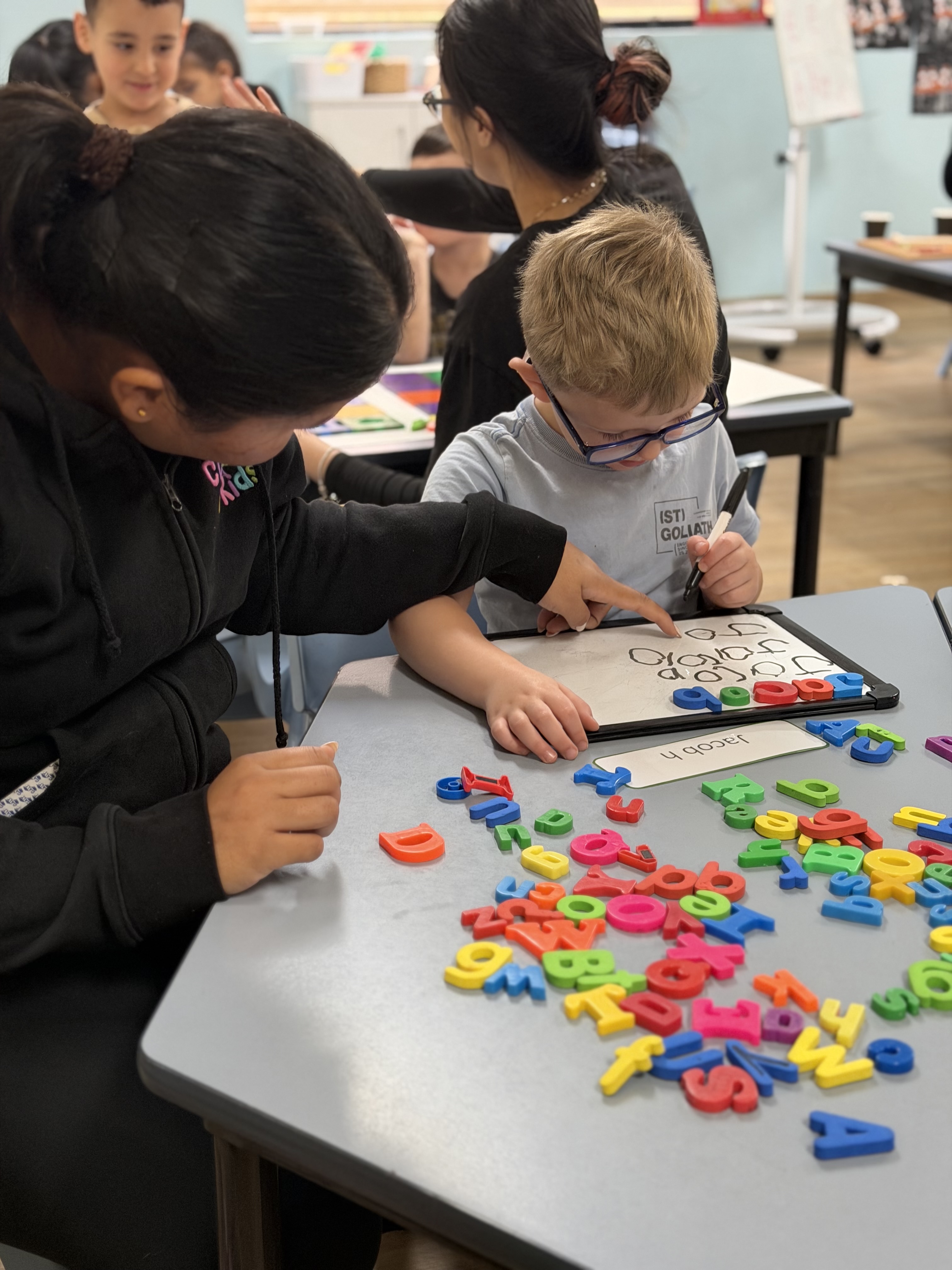 Educator assisting a preschool boy practicing name spelling with colorful magnetic letters on a whiteboard